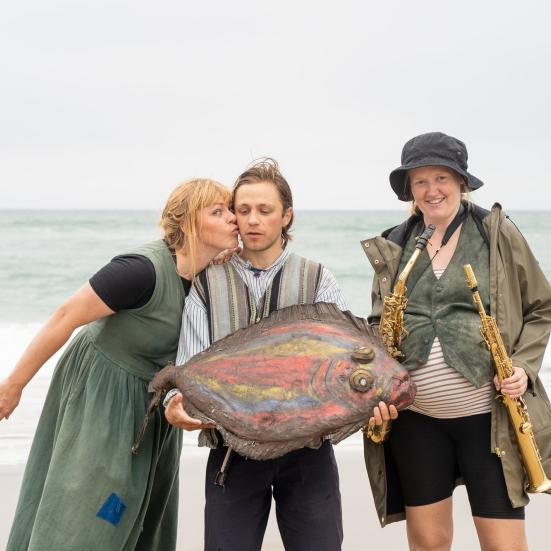 Tre skuespillere står på en strand ved Vesterhavet. I midten står en mand, som holder en farverig kunstig flynder. Kvinden til den ene side kysser ham på kinden. Kvinden på den anden side holder et blæseinstrument i hver hånd.  