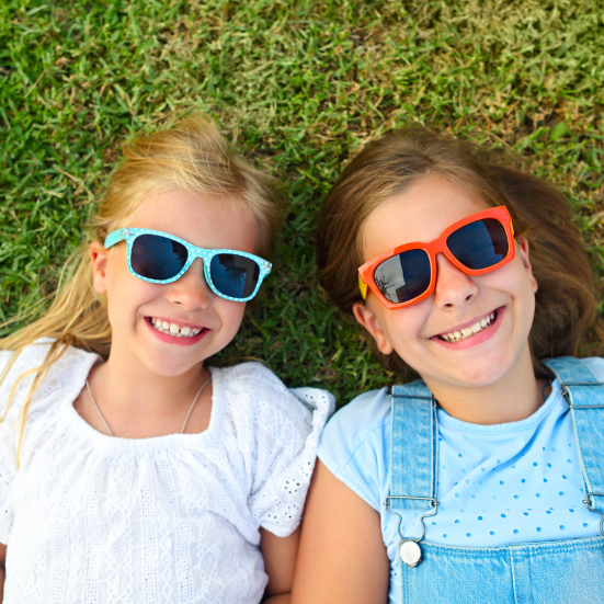 To smilende piger ligger i græsset i sommersolen, og de har solbriller på.