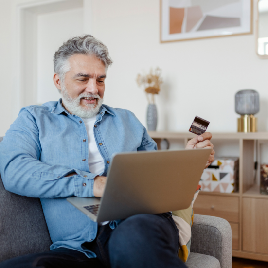 En ældre herre sidder i sin sofa med en computer på skødet og et kreditkort i hånden. I baggrunden ses resten af en hyggelig stue.