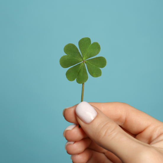 En hånd med lys neglelak på neglene holder et firkløver op mod en turkis baggrund.