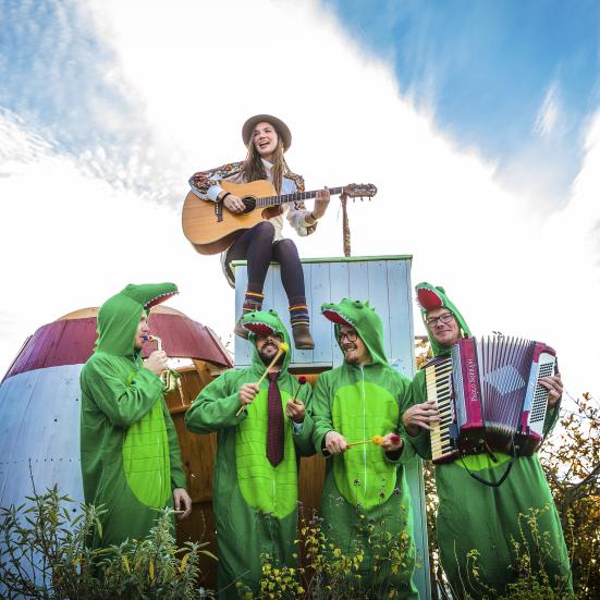 En kvinde med en guitar sidder på et skur. Under hende står fire mænd klædt ud som krokodiller og med instrumenter i hænderne. 