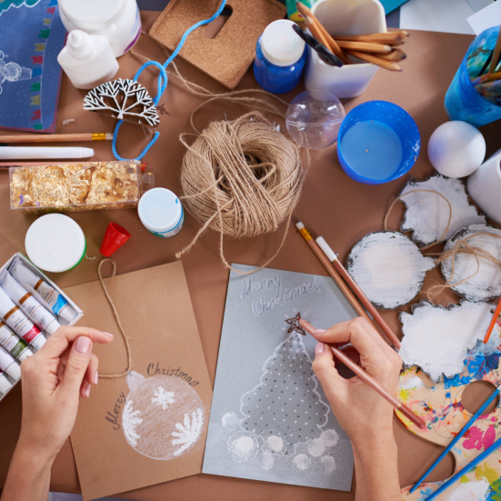 Et bord fuld af kreative materialer og to hænder, der er i gang med at lave et julekort.