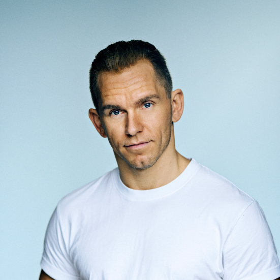 En mand står i en hvid t-shirt på en blå baggrund og kigger ind i kameraet.