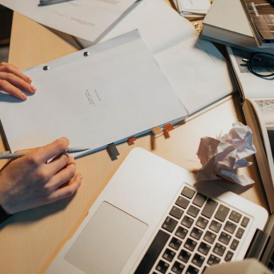 Hænder ved et skrivebord med manuskriptsider, bøger og en bærbar computer.