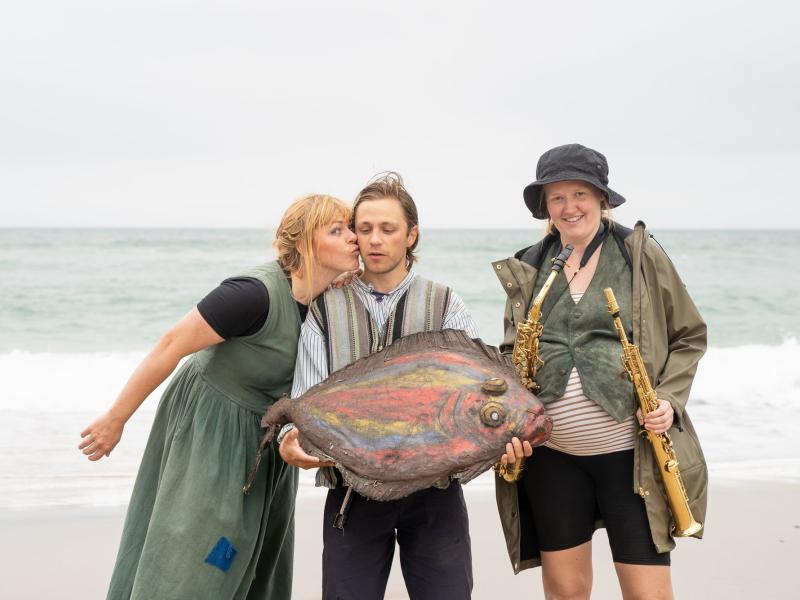 Tre skuespillere står på en strand ved Vesterhavet. I midten står en mand, som holder en farverig kunstig flynder. Kvinden til den ene side kysser ham på kinden. Kvinden på den anden side holder et blæseinstrument i hver hånd.  