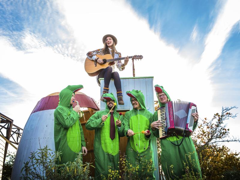 En kvinde med en guitar sidder på et skur. Under hende står fire mænd klædt ud som krokodiller og med instrumenter i hænderne. 