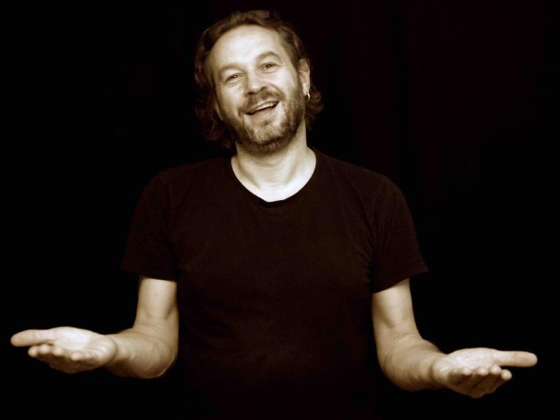 En mand i sort trøje og på en sort baggrund står med armene strakt ud til siden i færd med at fortælle.