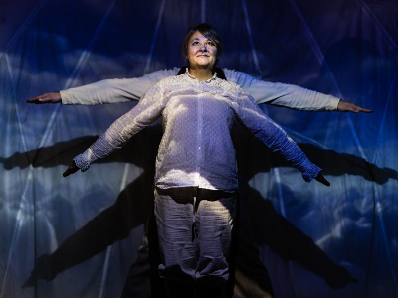 To mennesker i en teateropsætning med lys, den ene står forrest med armene spredt ud til siderne, den anden står bagerst, så man kun kan se vedkommendes arme. 