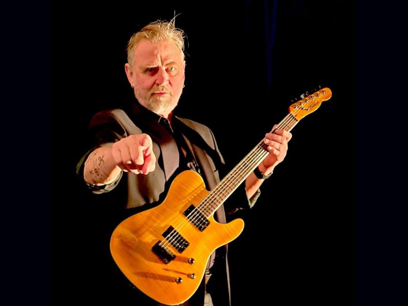 En mand står med en elektrisk guitar og peger ud mod beskueren. 