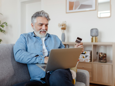 En ældre herre sidder i sin sofa med en computer på skødet og et kreditkort i hånden. I baggrunden ses resten af en hyggelig stue.