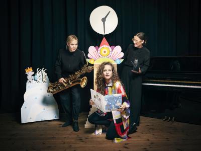 Tre kvinder sidder i en teaterkulisse. Den ene med en bog, den anden med en saxofon og den tredje med et lille instrument. 