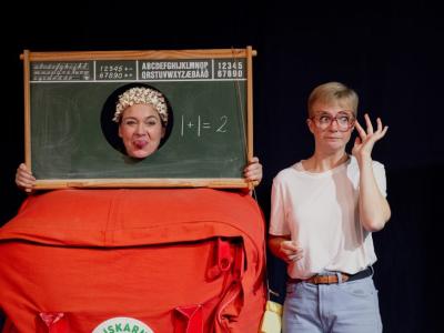 En kvinde står bag en stor skoletaske med en tavle, hvor der er hul til hovedet. Hun har en paryk på af popcorn. Ved siden af hende står en kvinde med store briller. 