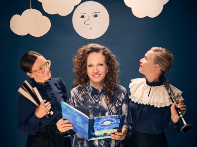 Tre kvinder i en teaterkulisse med en måne og skyer. Den ene kvinde holder en billedbog.