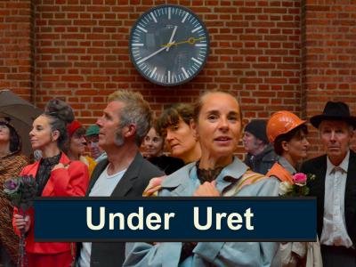 En masse mennesker på en togstation. De står under et stort ur. 