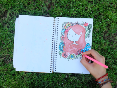 Et foto af en opslået notesbog, hvor man ser en hånd, der tegner et pigeansigt.