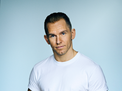 En mand står i en hvid t-shirt på en blå baggrund og kigger ind i kameraet.