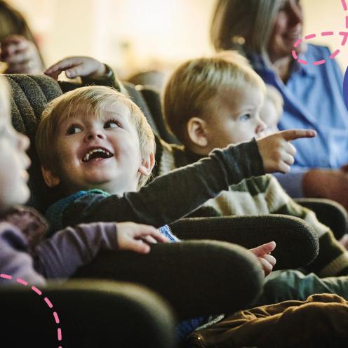 En flok små børn sidder i bløde stole omgivet af andre børn og voksne. Det ene barn peger begejstret op imod noget.