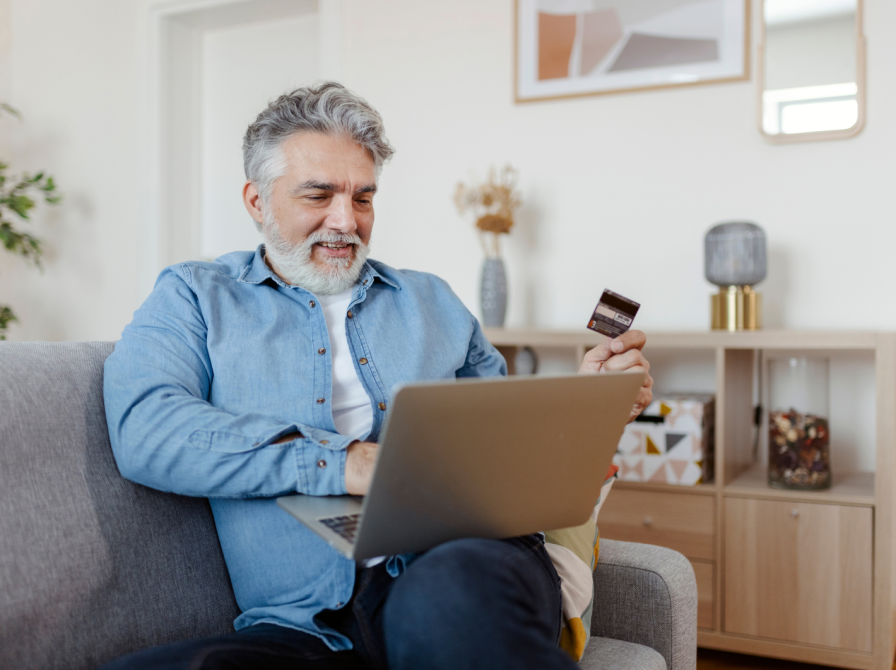 En ældre herre sidder i sin sofa med en computer på skødet og et kreditkort i hånden. I baggrunden ses resten af en hyggelig stue.