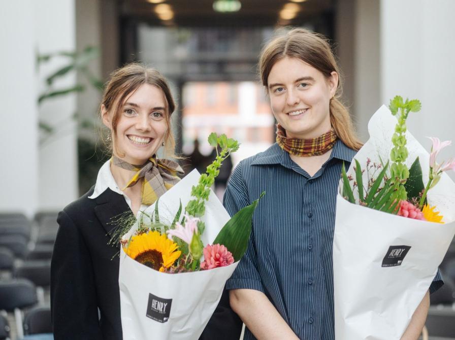 De to smilende prisvindere Helene Franch og Feline Kamber holder blomsterbuketter foran rækker af stole.